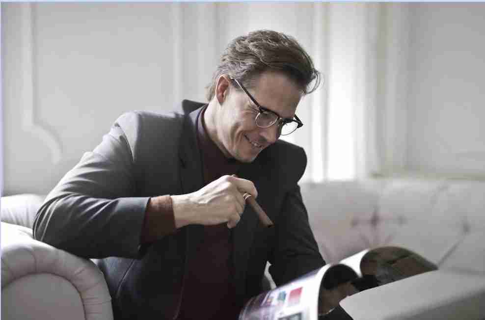 A man in a suit and glasses is smiling and gesturing triumphantly while looking at a magazine.